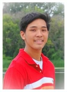 A young man wearing a red shirt with white and orange stripes, smiling while outdoors.